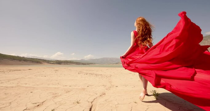 Young sexy elegant beautiful woman girl red flowing fluttering dress walks through desert. Gignger white skin model move sand hair flying wind. High-quality 4k horizontal footage slow motion