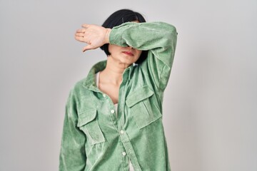 Young asian woman standing over white background covering eyes with arm, looking serious and sad....