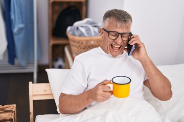 Middle age grey-haired man talking on smartphone drinking coffee at bedroom