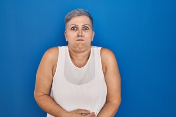 Middle age caucasian woman standing over blue background puffing cheeks with funny face. mouth...
