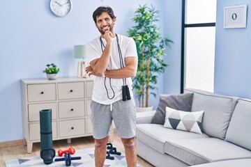 Handsome latin man wearing sportswear at home looking confident at the camera smiling with crossed arms and hand raised on chin. thinking positive.