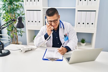 Young latin man doctor talking on telephone writing on document at clinic