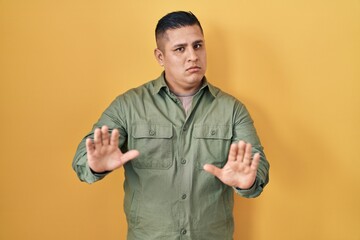 Hispanic young man standing over yellow background moving away hands palms showing refusal and...