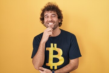 Hispanic young man wearing bitcoin t shirt looking confident at the camera smiling with crossed arms and hand raised on chin. thinking positive.