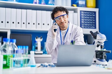 Young hispanic man scientist using laptop talking on smartphone at laboratory