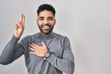 Hispanic man with beard standing over white background smiling swearing with hand on chest and...