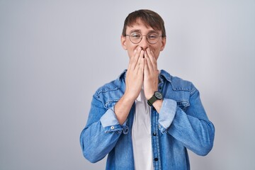 Caucasian blond man standing wearing glasses laughing and embarrassed giggle covering mouth with...