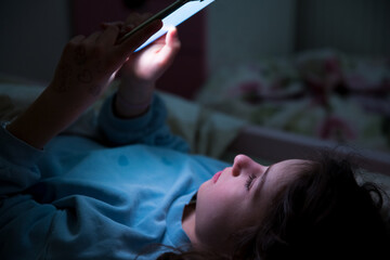 A child using smart phone lying in bed late at night, playing games, watching videos online,...