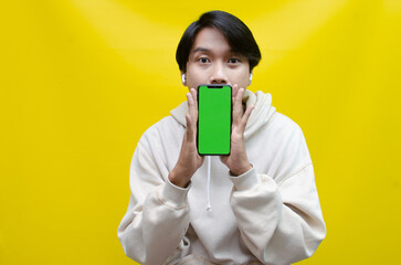 Asian man in beige hoodie using earphones, raising fist and showing his screen phone with green screen template best for advertisment. man showing mock up of phone.