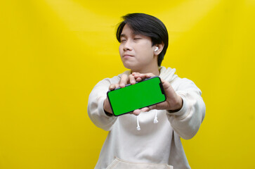 Asian man in beige hoodie using earphones, raising fist and showing his screen phone with green screen template best for advertisment. man showing mock up of phone.