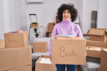 Young brunette woman with curly hair moving to a new home holding cardboard box celebrating crazy and amazed for success with open eyes screaming excited.