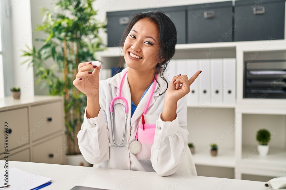 Poster young asian woman wearing doctor uniform and stethoscope smiling happy pointing with hand and finger