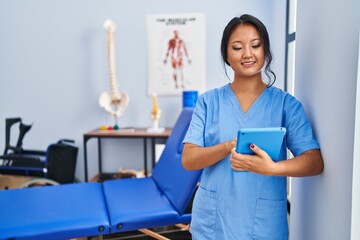 Young chinese woman wearing physiotherapist uniform using touchpad at rehab clinic