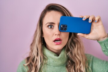 Young caucasian woman covering eye with smartphone in shock face, looking skeptical and sarcastic, surprised with open mouth