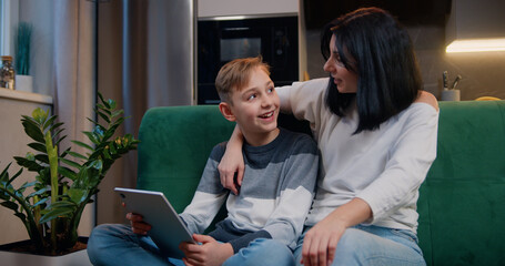Boy and woman playing in game at tablet. Son and mother sitting on sofa, sharing tablet pc, networking and discussing social media content online while relaxing indoors.