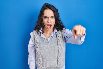 Young brunette woman standing over blue background pointing displeased and frustrated to the...