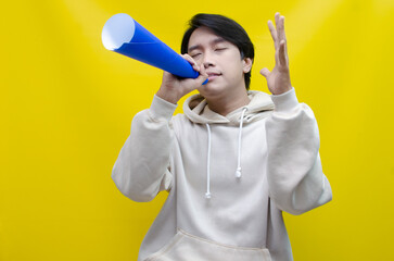 An ecstatic young Asian man in a cream hoodie whispering and shouting through a paper megaphone giving an announcement. asian man dancing and singing happily using paper as a megaphone.
