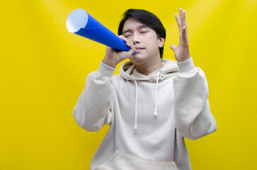 An ecstatic young Asian man in a cream hoodie whispering and shouting through a paper megaphone...