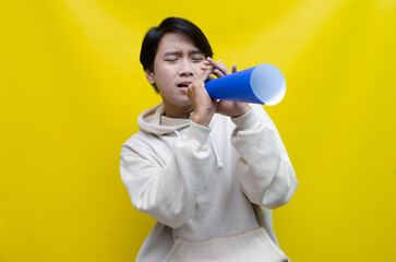 An ecstatic young Asian man in a cream hoodie whispering and shouting through a paper megaphone giving an announcement. asian man dancing and singing happily using paper as a megaphone.
