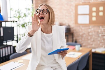 Young caucasian woman working at the office wearing glasses shouting and screaming loud to side with hand on mouth. communication concept.