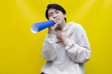 An ecstatic young Asian man in a cream hoodie whispering and shouting through a paper megaphone...