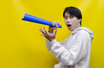 An ecstatic young Asian man in a cream hoodie whispering and shouting through a paper megaphone...