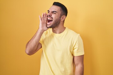 Young hispanic man standing over yellow background shouting and screaming loud to side with hand on mouth. communication concept.