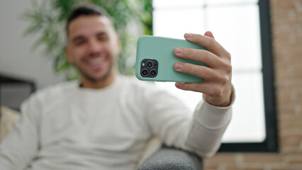 Young hispanic man watching video on smartphone sitting on sofa at home