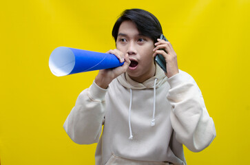 expressive Asian man holding phone while shouting to paper megaphone to give an offering and...