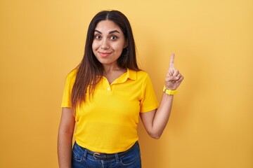 Young arab woman standing over yellow background showing and pointing up with finger number one while smiling confident and happy.