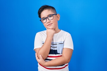 Young hispanic kid standing over blue background with hand on chin thinking about question, pensive expression. smiling with thoughtful face. doubt concept.