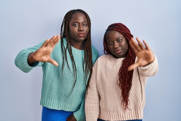Two african woman standing over blue background doing frame using hands palms and fingers, camera perspective - Powered by Adobe