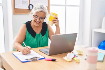 Middle age grey-haired woman business worker writing on document drinking coffee at office