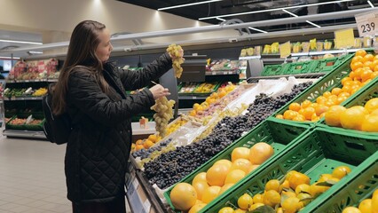 A young woman buys grapes in a supermarket. A woman at a fruit stand chooses grapes. The lady is buying fruit for dessert. Supermarket shopping concept