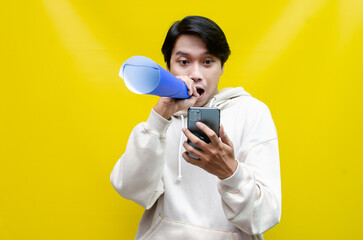 expressive Asian man holding phone while shouting to paper megaphone to give an offering and...