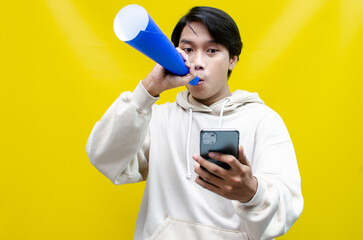 expressive Asian man holding phone while shouting to paper megaphone to give an offering and...