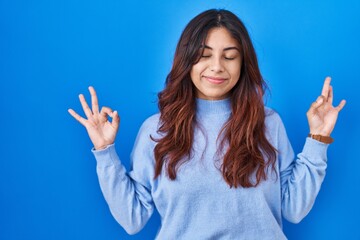 Hispanic young woman standing over blue background relaxed and smiling with eyes closed doing meditation gesture with fingers. yoga concept.
