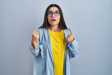 Young hispanic woman standing over blue background amazed and surprised looking up and pointing with fingers and raised arms.