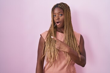 African american woman with braided hair standing over pink background surprised pointing with finger to the side, open mouth amazed expression.