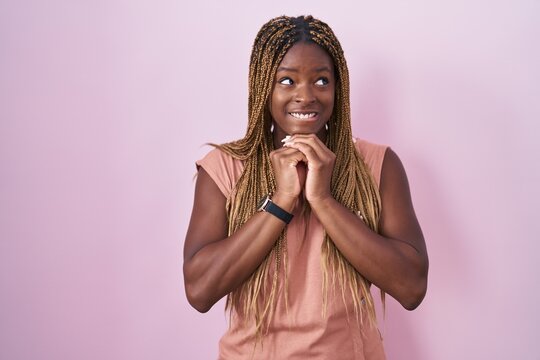 African american woman with braided hair standing over pink background laughing nervous and excited with hands on chin looking to the side