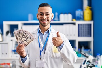 Young hispanic man working at scientist laboratory holding money smiling happy and positive, thumb up doing excellent and approval sign