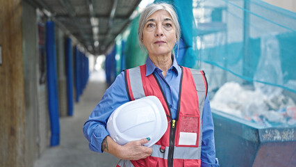 Middle age grey-haired woman builder standing with relaxed expression holding hardhat at street