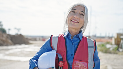 Middle age grey-haired woman builder smiling confident holding hardhat at street