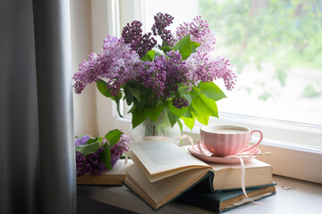 A cup of freshly brewed coffee with heart-shaped foam and spring lilac flowers, open books. Spring still life.