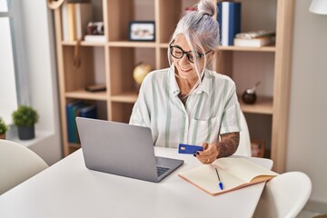 Middle age grey-haired woman using laptop and credit card sitting on table at home