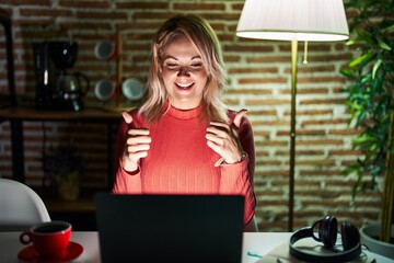 Fototapeta na wymiar Blonde woman using laptop at night at home success sign doing positive gesture with hand, thumbs up smiling and happy. cheerful expression and winner gesture.