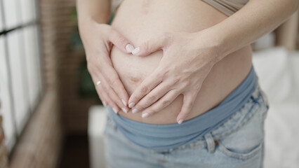 Young pregnant woman doing heart gesture with hands on belly at bedroom