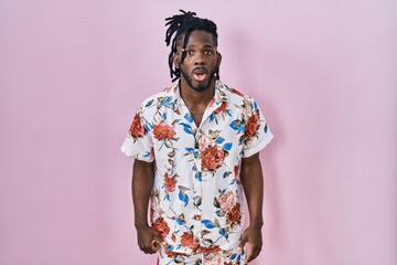 African man with dreadlocks wearing summer shirt over pink background scared and amazed with open...