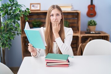 Young caucasian woman reading books at home covering mouth with hand, shocked and afraid for mistake. surprised expression