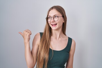 Young caucasian woman standing over white background smiling with happy face looking and pointing to the side with thumb up.